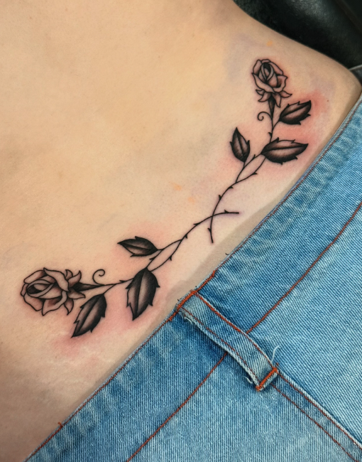 A black and gray floral tattoo of a rose and leaves on the person's side hip, partially covered by blue jeans.