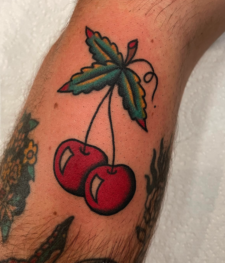 Tattoo of two red cherries with green and yellow leaves on a person's arm.