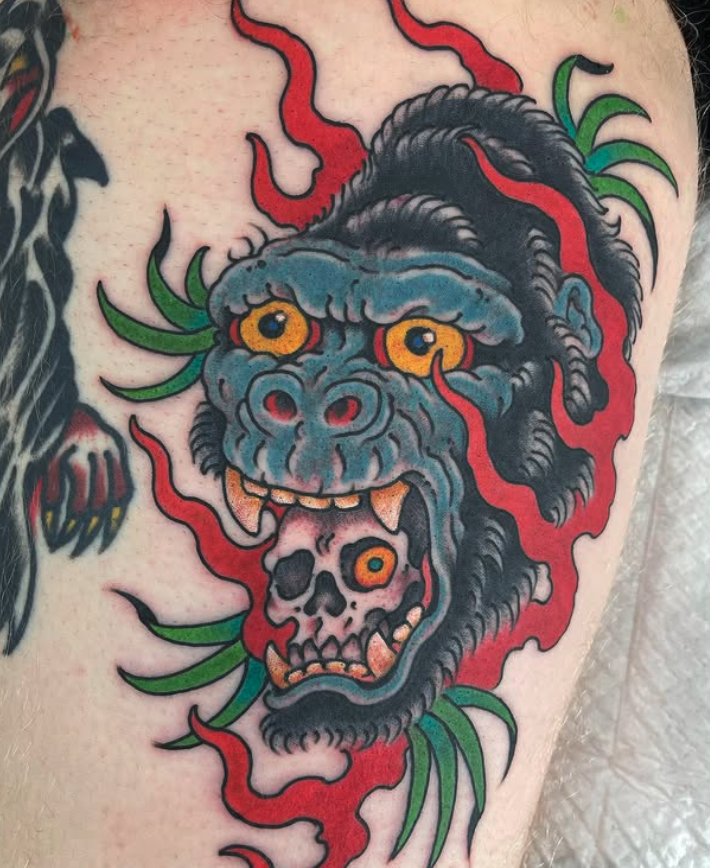 Tattoo of a mythical creature with a blue face, yellow eyes, and sharp teeth, holding a skull in its mouth, surrounded by red flames and green leaves.