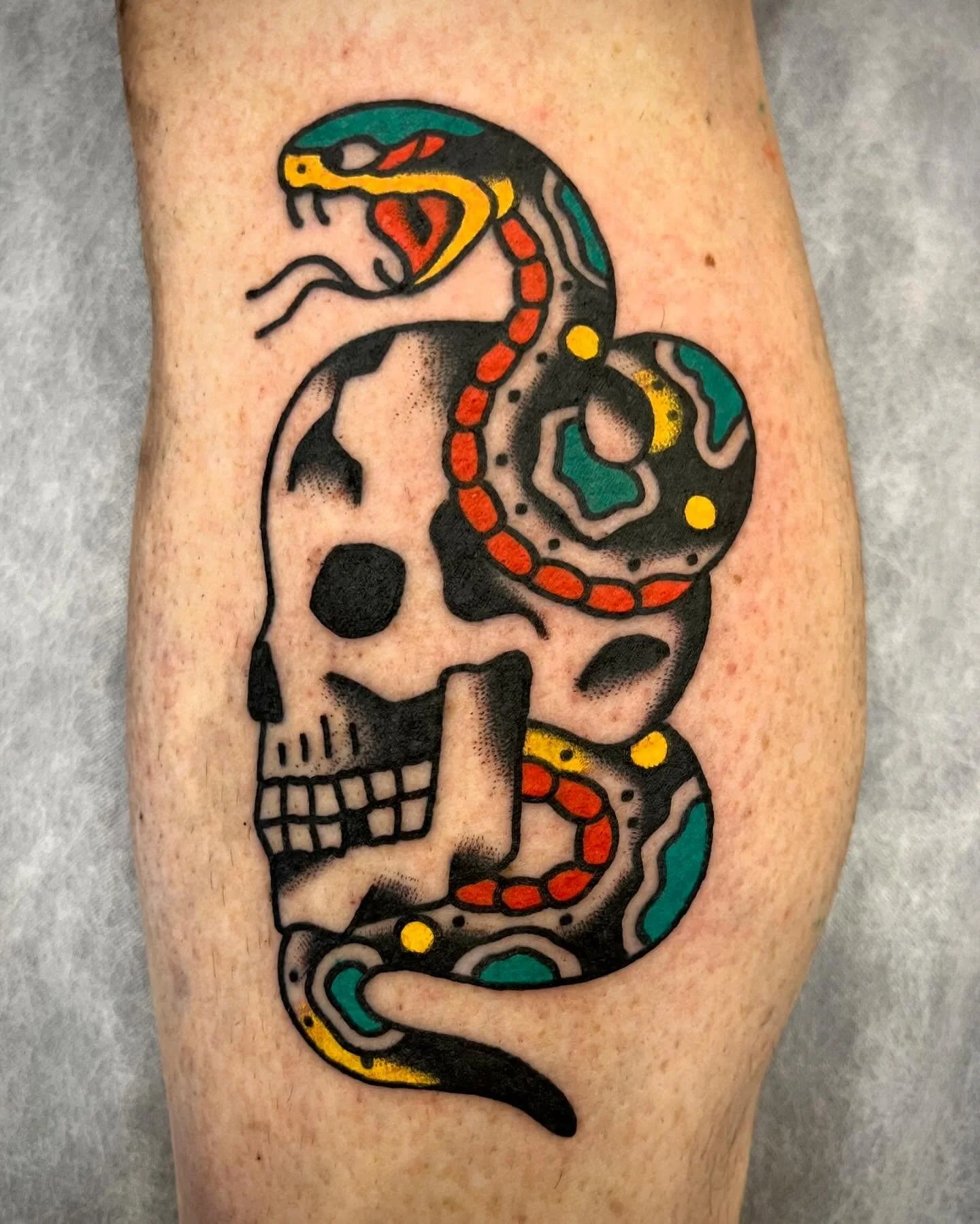 Tattoo of a skull with a snake wrapped around it, featuring black outlines, colorful accents in red, yellow, teal, and gray.