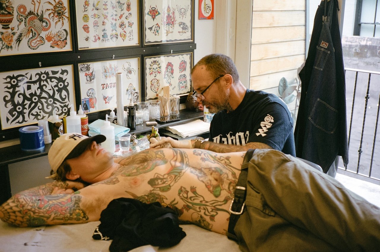 A man with tattoos lying on an adjacent table, receiving tattoo work from a tattoo artist. The artist is wearing glasses and a black shirt, focused on his work. The room has artwork displayed on the wall and tattoo supplies on a counter.