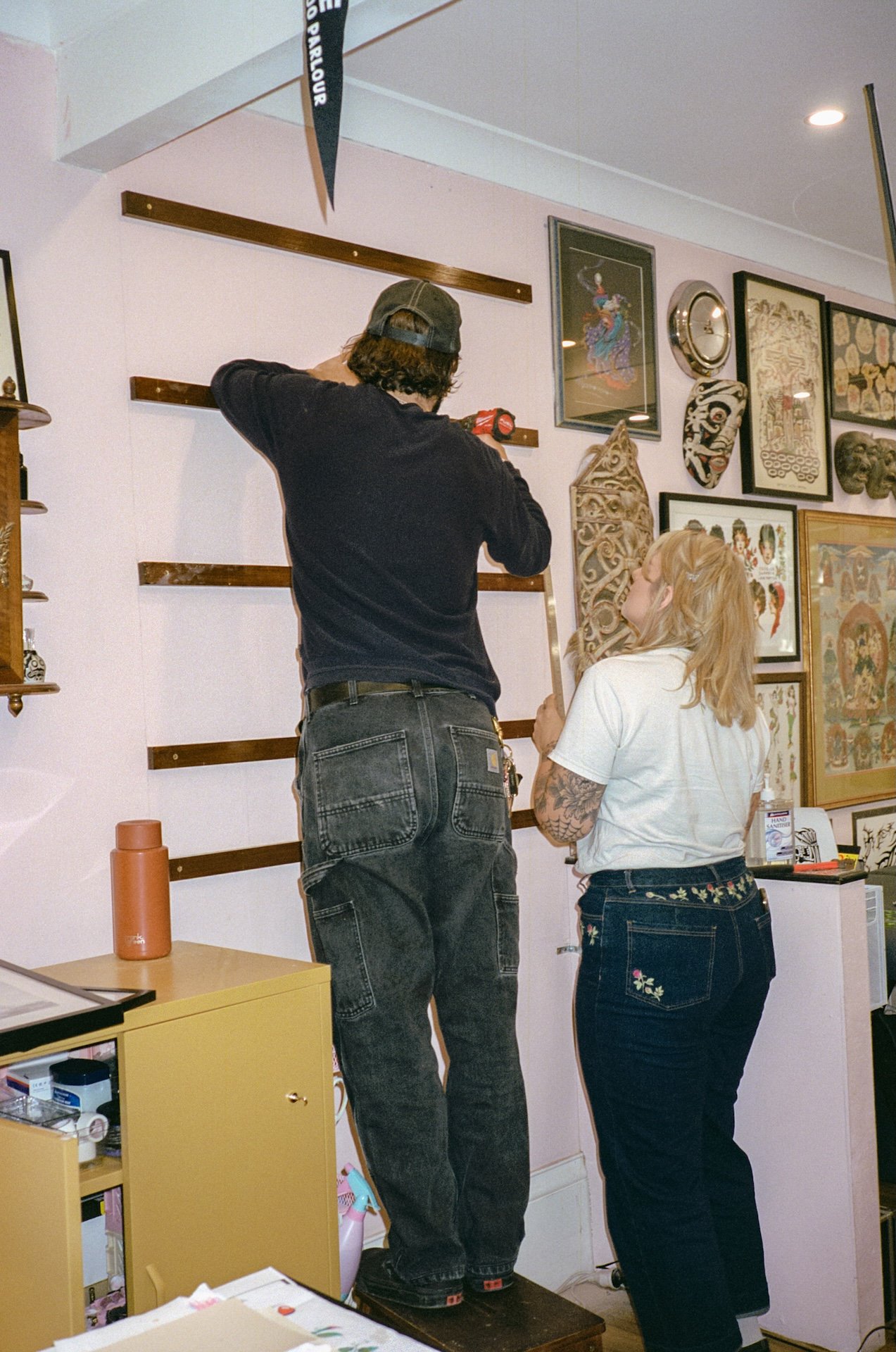 Two people working together to hang a gallery wall of assorted framed artworks and artifacts in a room.