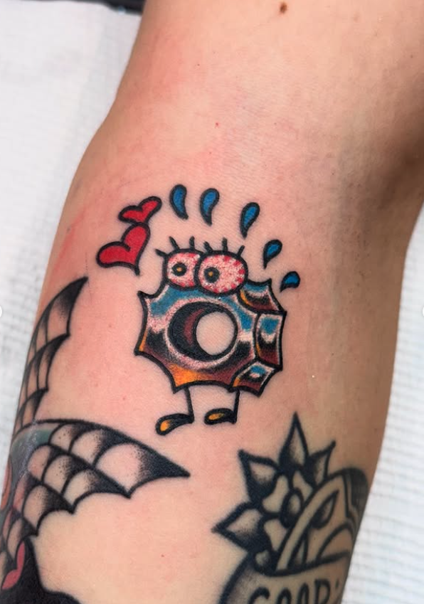 A colorful cartoon-style tattoo of a donut with eyes, a smiling mouth, and blue sprinkles, with a red heart and blue teardrops above it, on a person's arm.