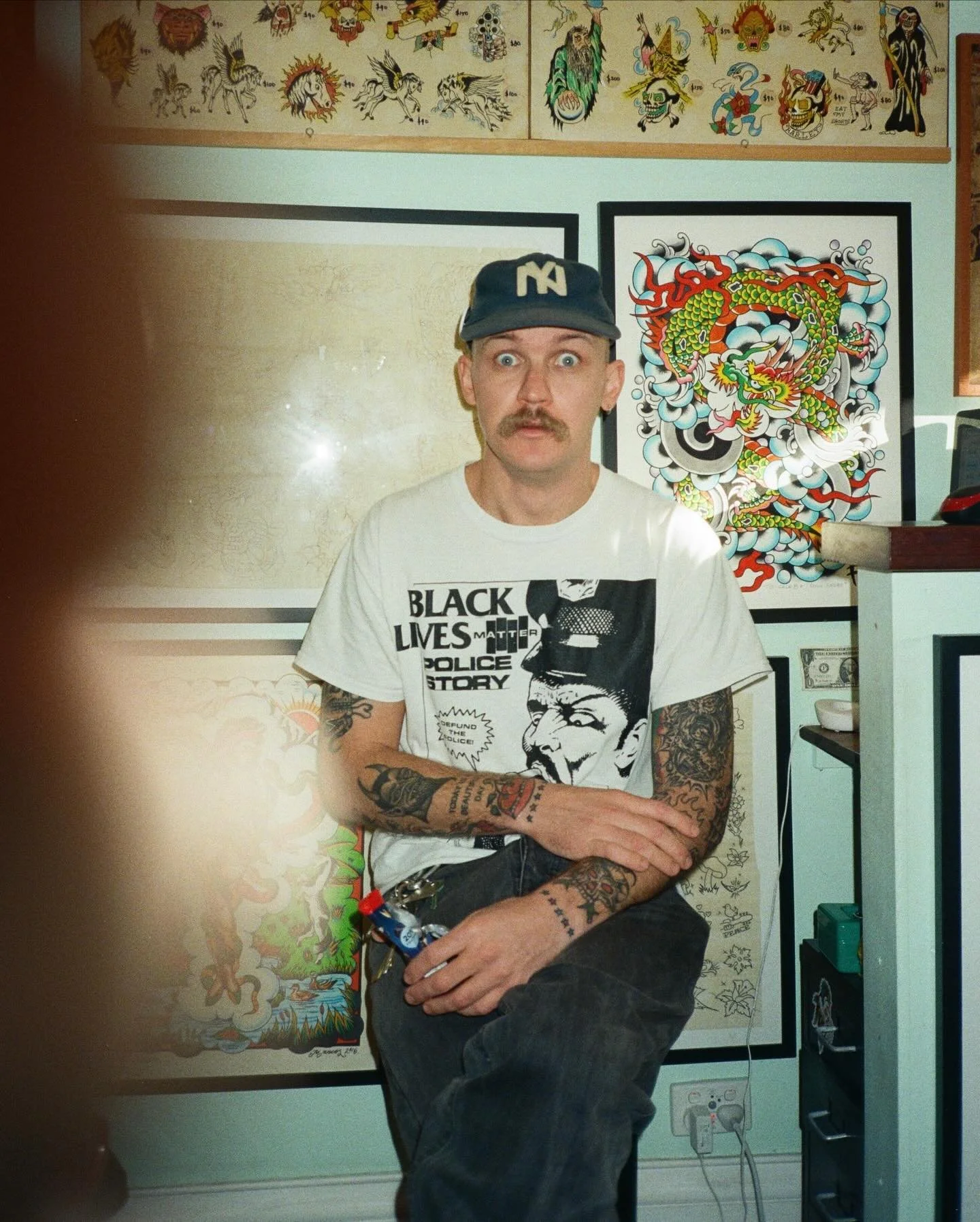 A man with tattoos, a mustache, wearing a graphic T-shirt and a baseball cap. He is sitting with one leg crossed over the other in front of a wall with tattoos and artwork, including a colorful dragon tattoo on the wall behind him.