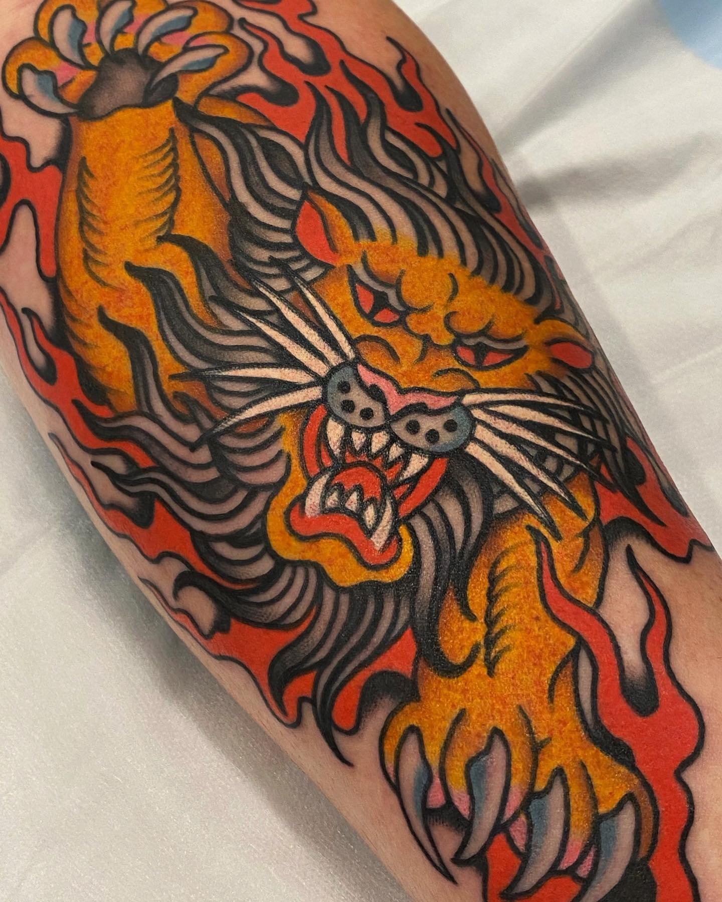 Colorful tattoo of a fierce tiger's face with sharp teeth, surrounded by flames and bold black lines.