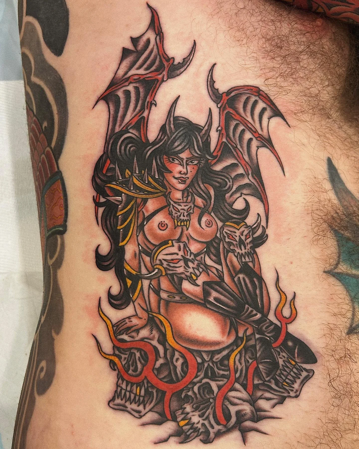 Tattoo of a female demon with black hair, wings, and armor, seated on a skull with fire around her.