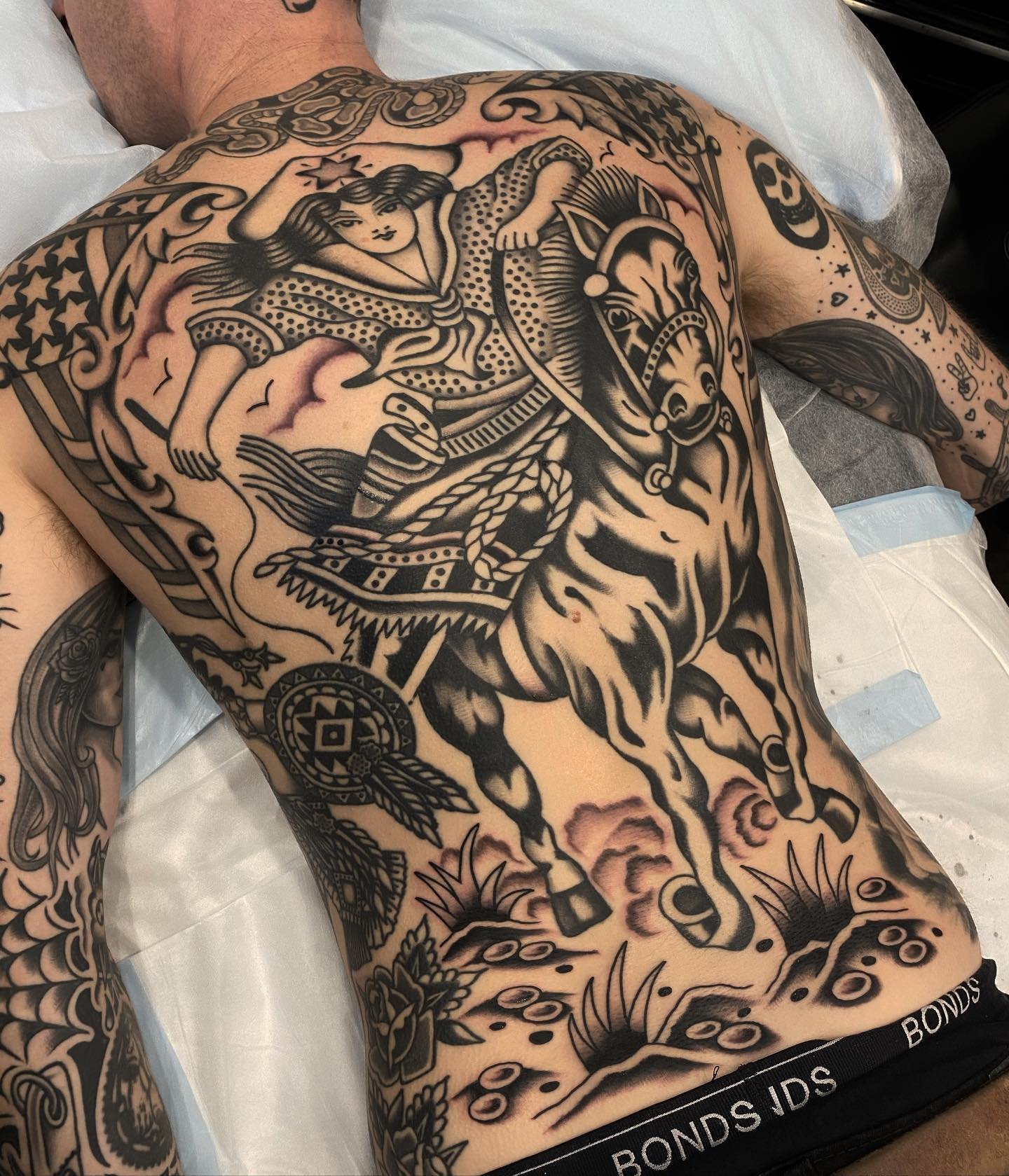 A person lying on a bed with a large black tattoo covering their chest, stomach, and part of their arms. The tattoo depicts a Native American warrior riding a horse, holding a spear, with decorative elements like feathers and geometric patterns.