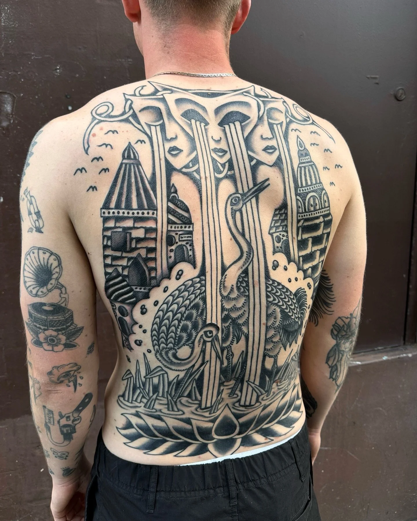 Back tattoo featuring female faces, buildings, a snake, a crane, and other symbolic elements in black ink.