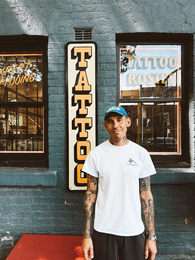 A man with tattoos on both arms and wearing a white t-shirt and blue cap standing outside a tattoo shop with a vertical sign that says 'TATTOO' and windows with signs for tattoo and tattoo Rosie.