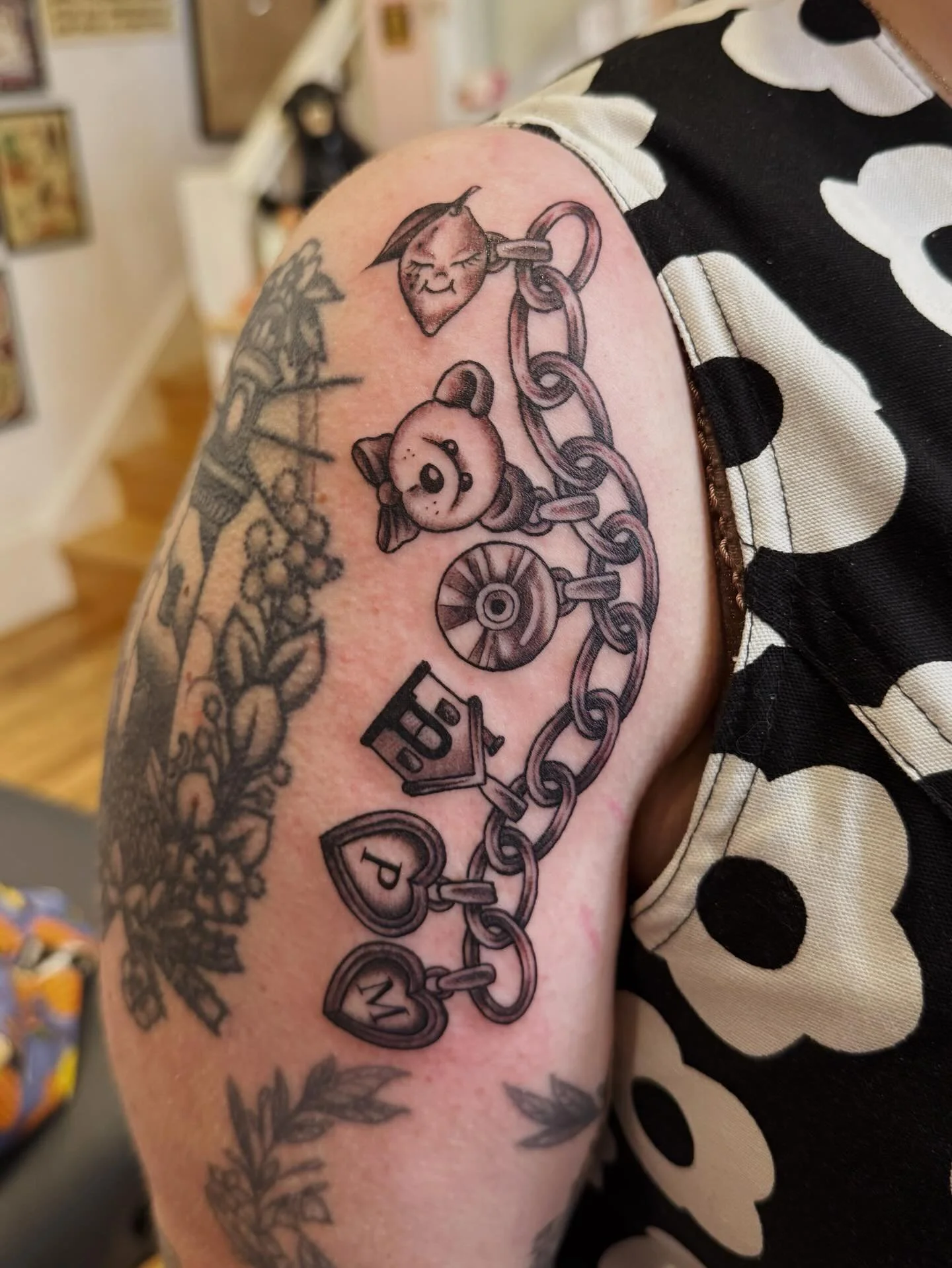 Tattoo of a chain with heart-shaped and round charms featuring letters, a panda, and a smiling face on a person's upper arm.