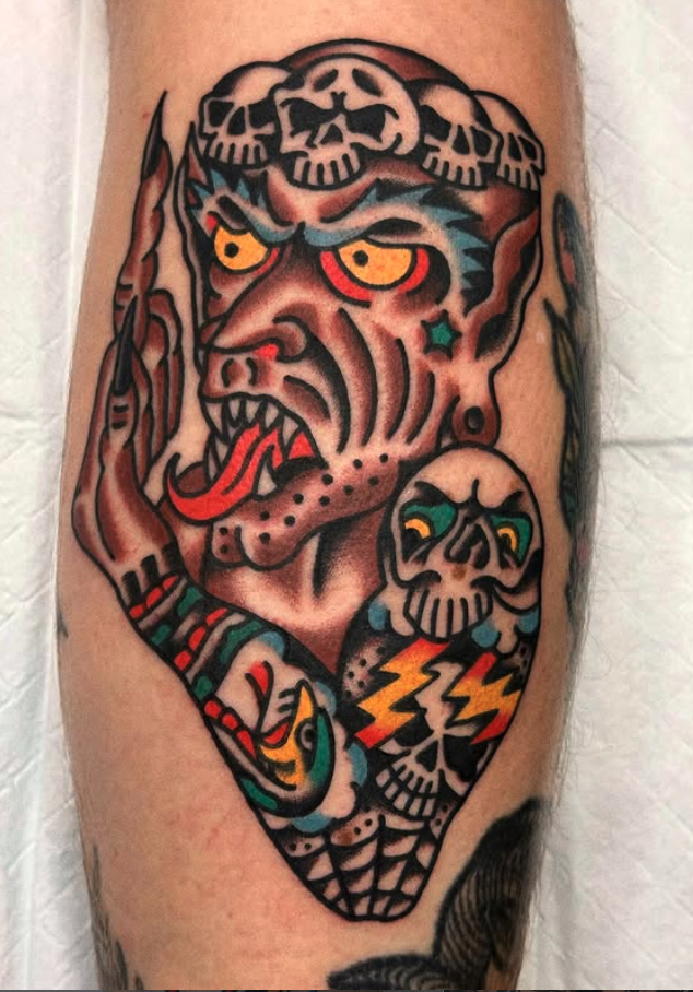 A colorful tattoo of a fierce, mythical creature with yellow eyes, a snarling face with tusks, and a crown of skulls. The creature has blue and black features, with a skull and lightning bolt design on its arm.