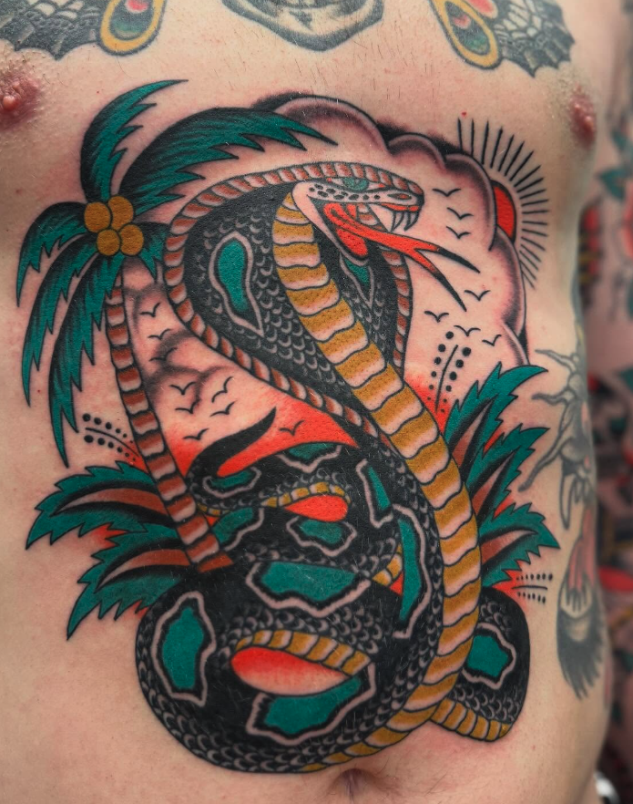 Tattoo of a coiled snake with open mouth, surrounded by palm trees, birds, and a setting sun.