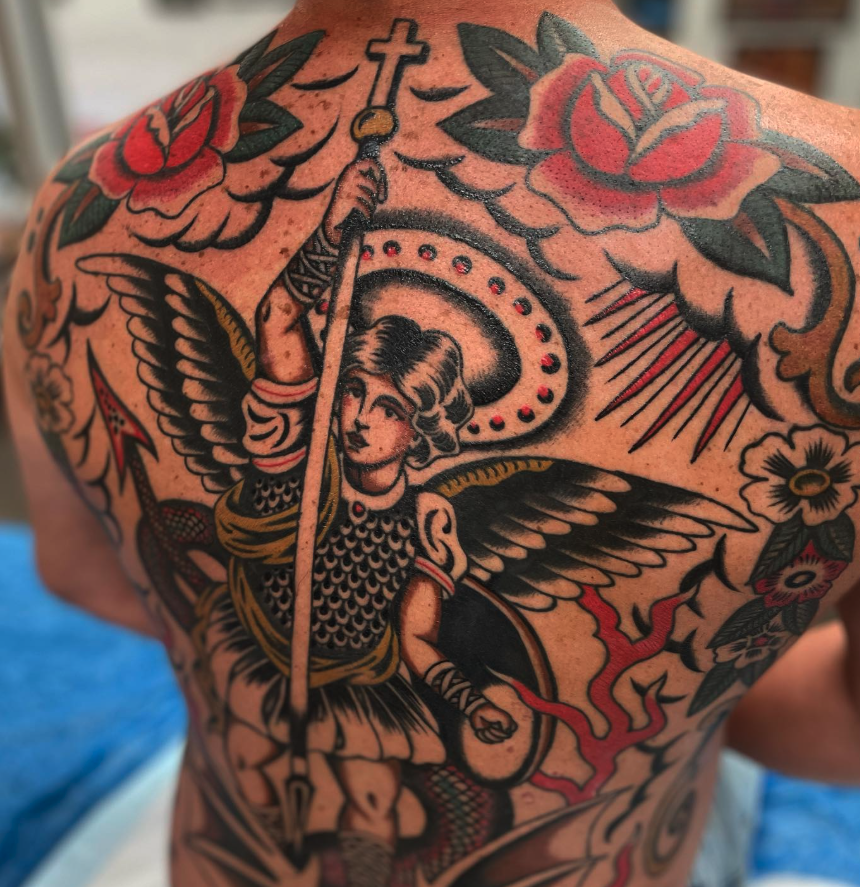 Tattoo of an angel with wings, holding a spear with a cross on top, surrounded by roses, clouds, and decorative elements on a person's back.