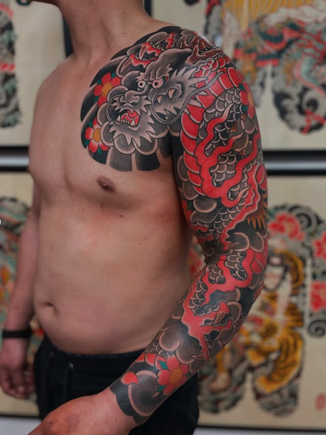 Tattoo of a fierce, traditional Asian dragon with gray, red, and black scales, on a man's upper arm and shoulder, with colorful flowers nearby.