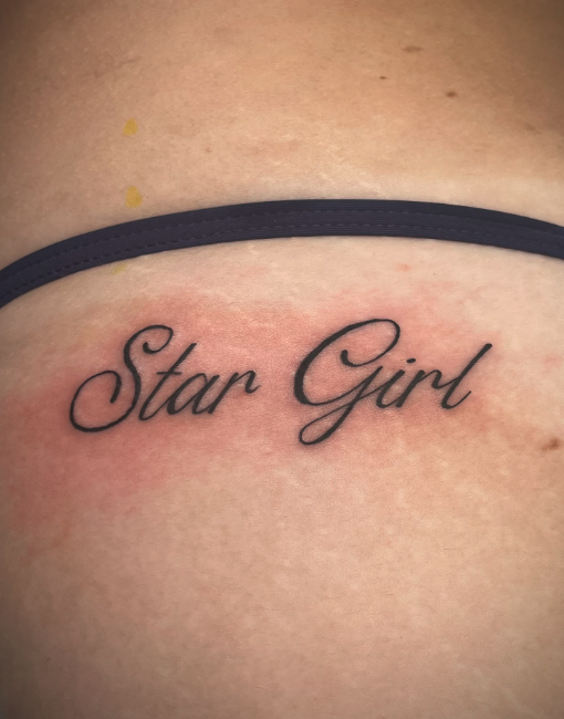 Tattoo on skin reading 'Star Girl' in cursive font beneath a dark strap, with some skin redness around the tattoo.