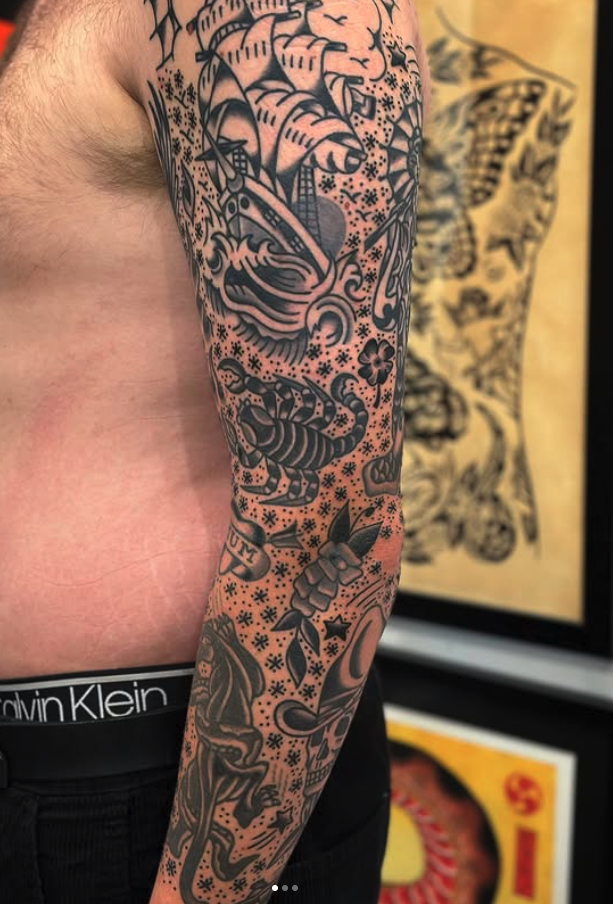 Tattoo sleeve on person's arm with traditional American tattoo designs including a ship, crab, scorpion, compass rose, dagger, skull, and various decorative elements.