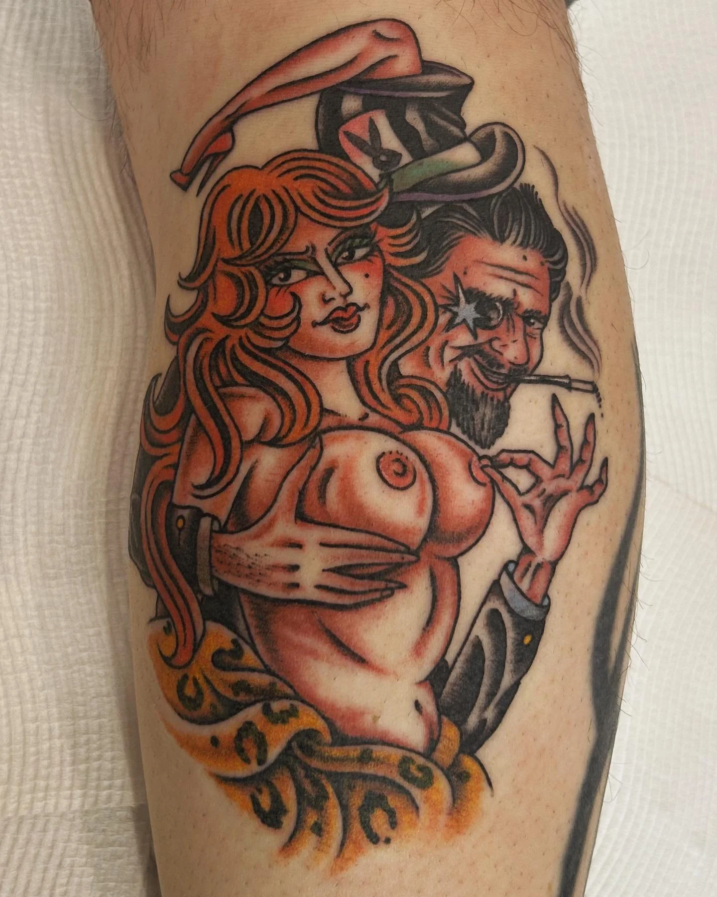 Tattoo of a woman with red hair and a man with a cigarette, both in a cartoon style, with the woman having exaggerated features and the man wearing a top hat and bow tie. The woman is topless, and there are decorative elements around them.