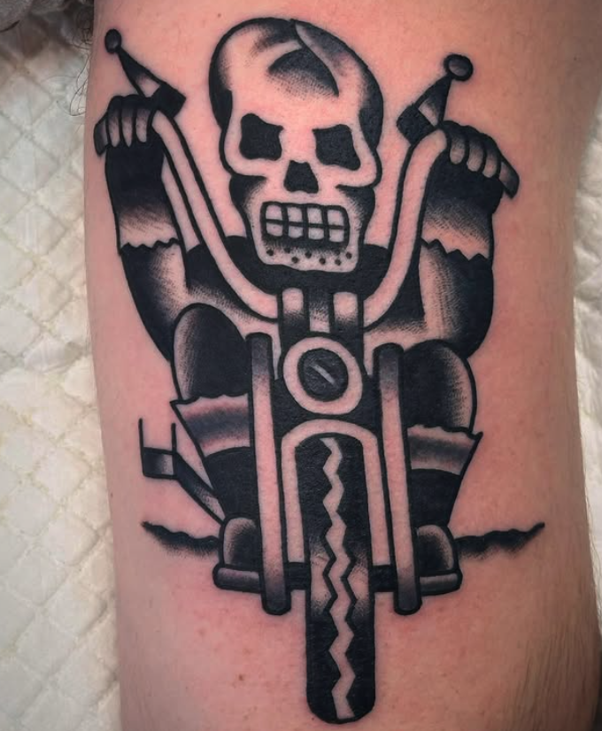 Tattoo of a skull riding a motorcycle with arms raised, detailed in black ink.