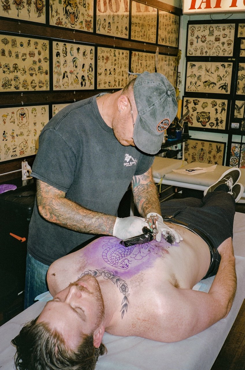 A tattoo artist is creating a large dragon tattoo on the chest of a shirtless man lying on a tattoo table, with tattoo flash art on the walls in the background.