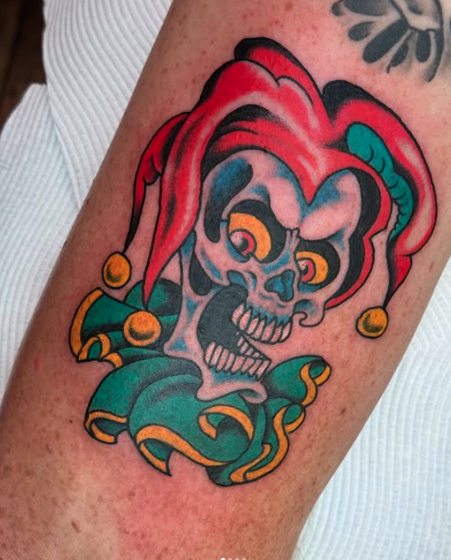Colorful tattoo of a grinning skull wearing a jester's hat, with swirling ribbon-like elements around it.