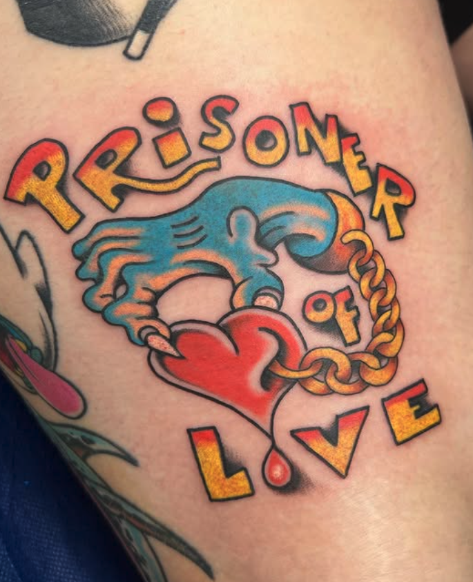 A colorful tattoo of a bleeding heart with a hand holding it, surrounded by the words 'Prisoner of Love' in bold, stylized letters.