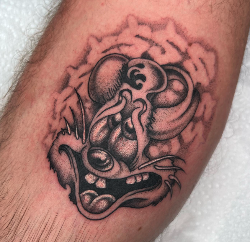 A black and gray tattoo of a creature with exaggerated features, including a large open mouth with sharp teeth, prominent eyes, and tentacle-like appendages.