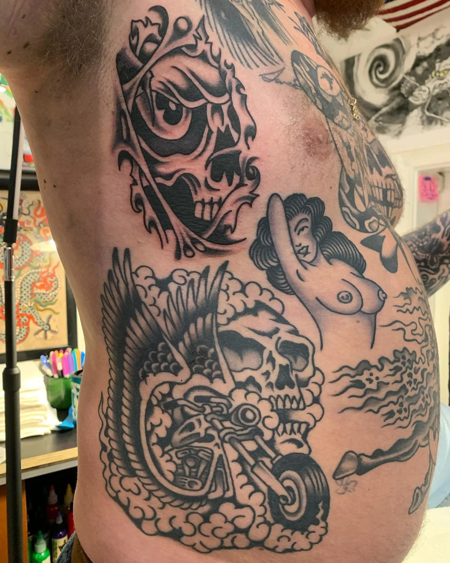 Tattoo of a skull, a woman with curly hair and nudity, a motorcycle, and a dragon in black ink on a person's side.
