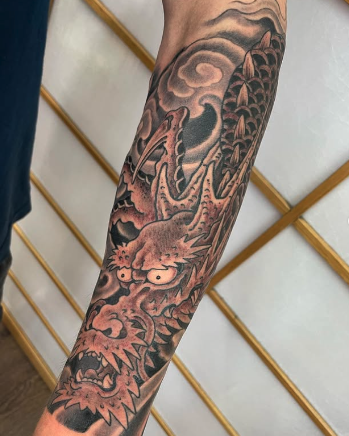 Black and gray tattoo of a fierce dragon with wide eyes and sharp teeth on a person's forearm, with clouds and flames in the background.