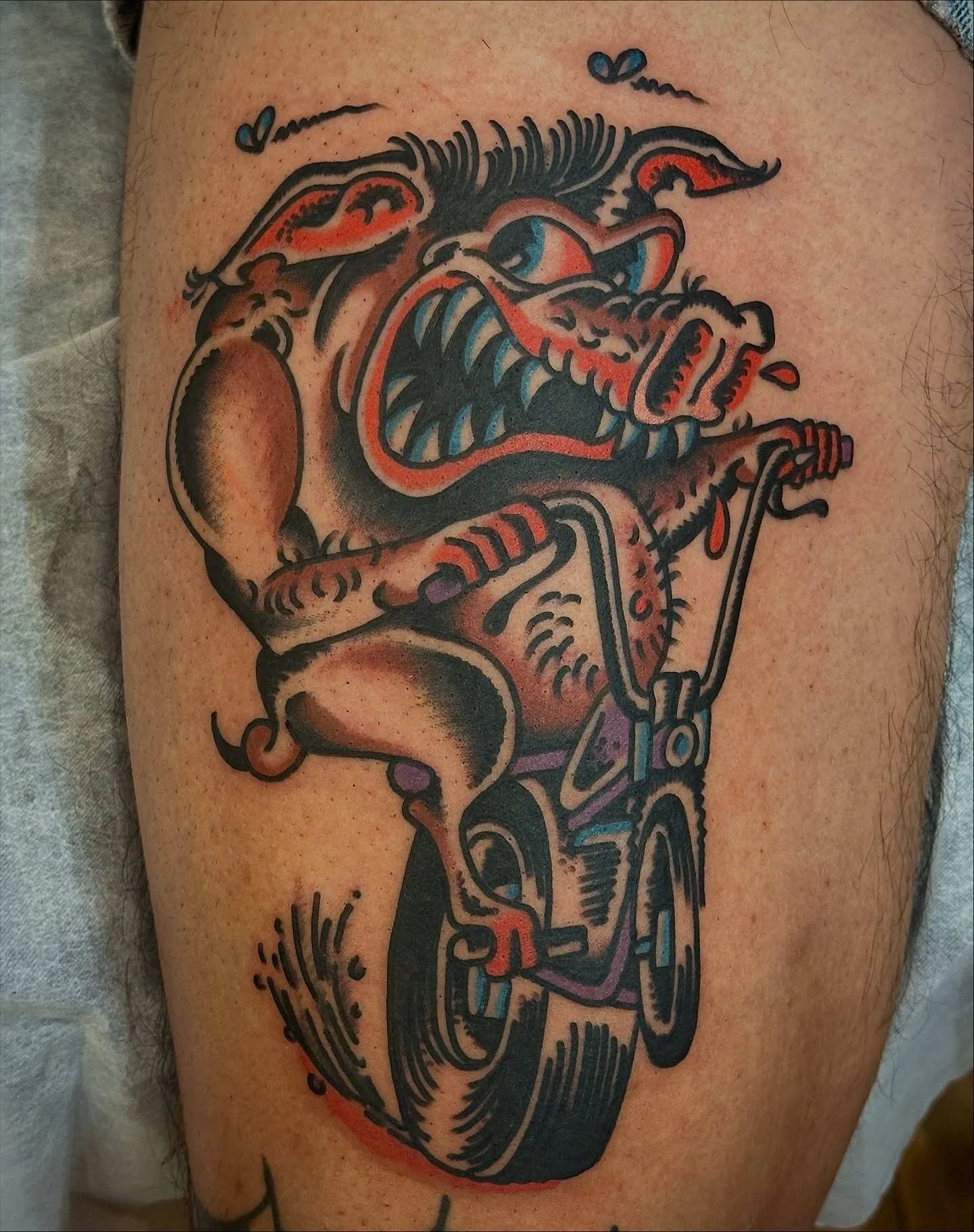 Colorful tattoo of a snarling dog with exaggerated features riding a bicycle, with a small red drip of blood near its mouth.