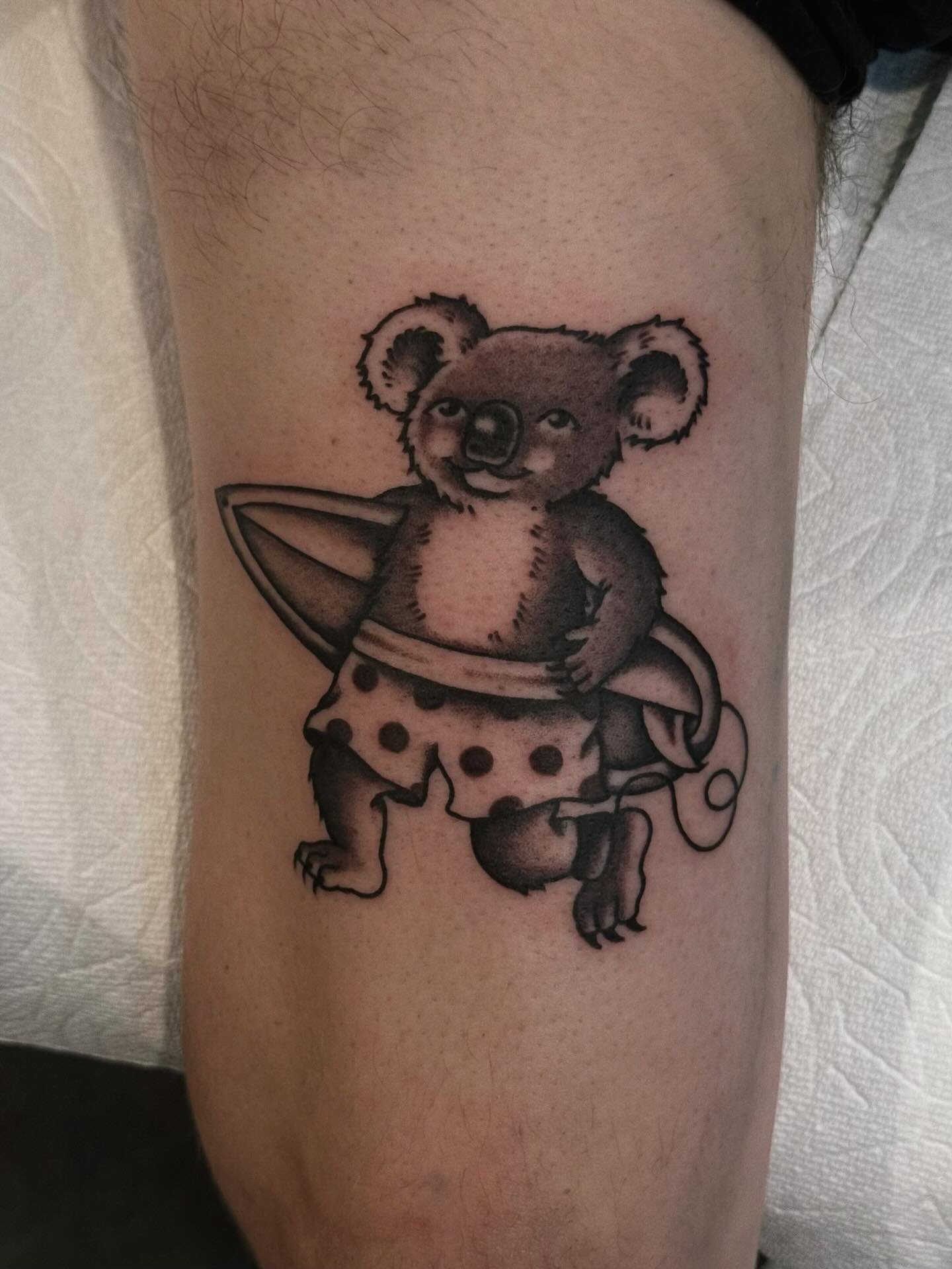 Tattoo of a koala bear with a surfboard, wearing polka-dotted swim trunks, on a person's leg.