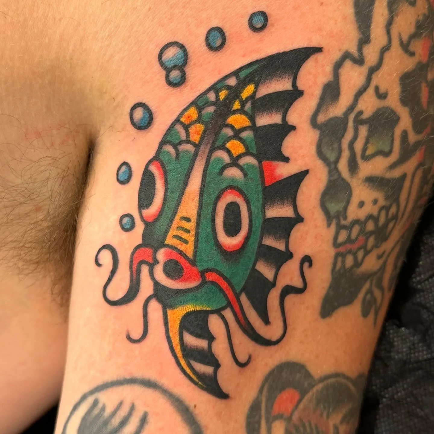 Colorful tattoo of a stylized fish with green, orange, and yellow accents, surrounded by bubbles, on a person's upper arm.