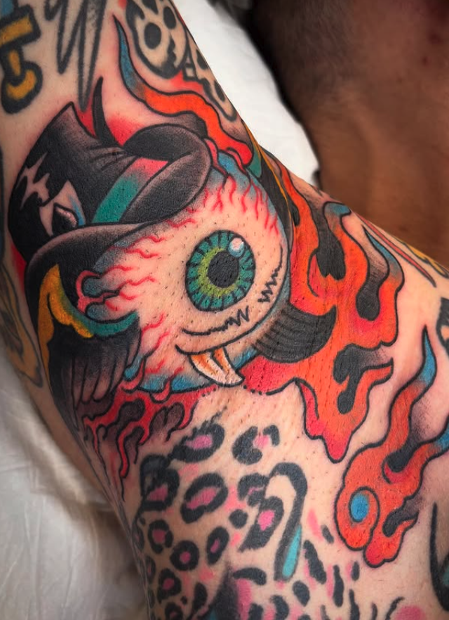 Tattoo of a cartoonish green-eyed eyeball with a tongue, surrounded by flames, on a person's arm.