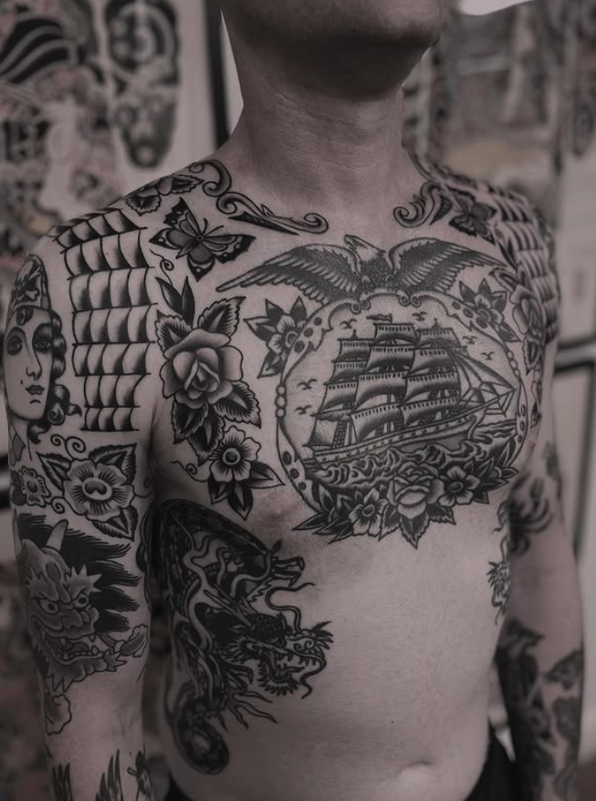 A person with a large collection of black and gray tattoos on their chest and arms, including a ship, an eagle, flowers, a face, and a dragon.