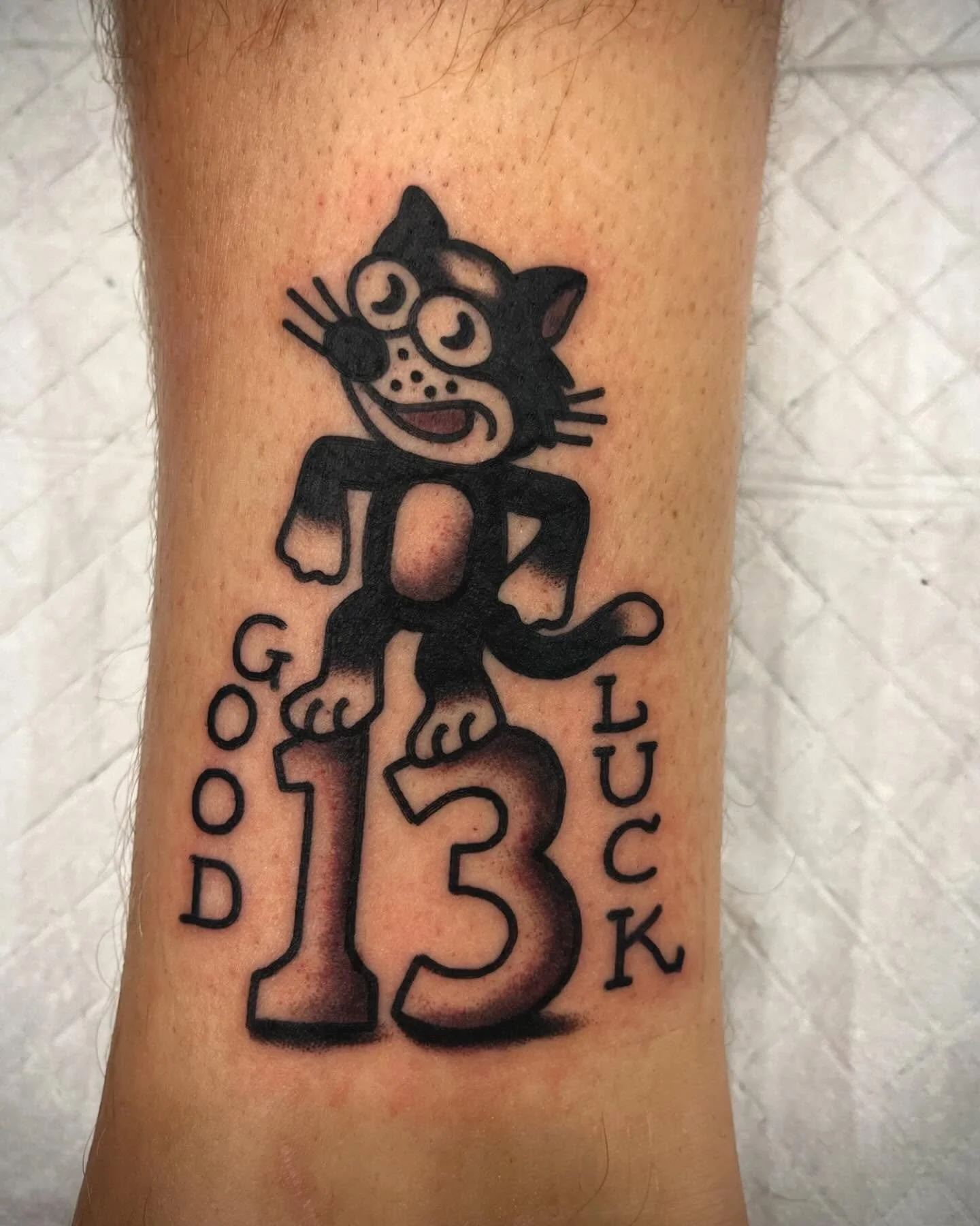 Tattoo of a cartoon cat with glasses, standing on a large number 13, with the words 'Good Luck' vertically on both sides.