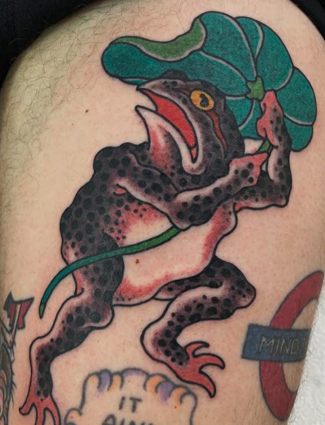 Colorful tattoo of a frog with a duck bill, holding a large green leaf.