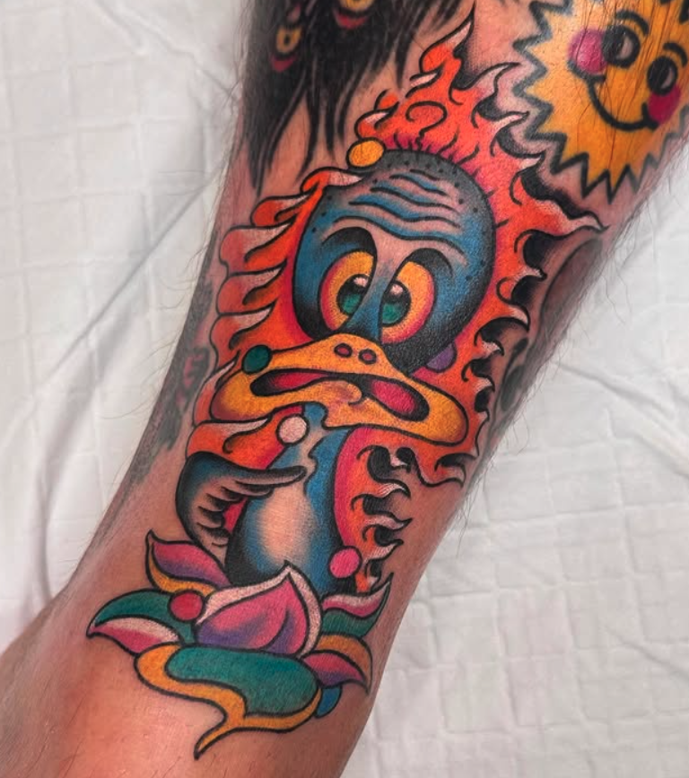 Colorful tattoo on a person's arm featuring a cartoonish bird with exaggerated features, surrounded by abstract flames and a smiling sun.
