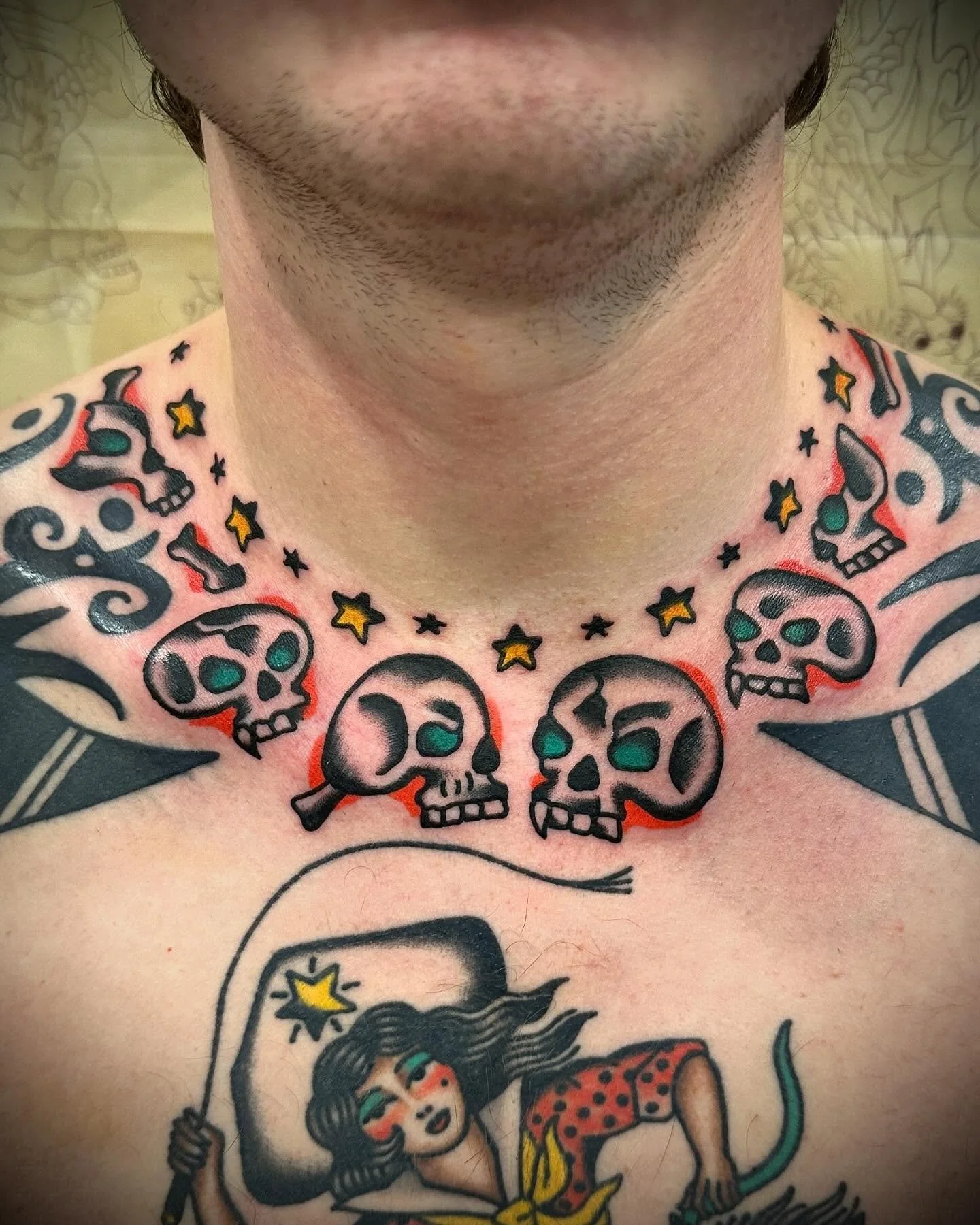 Tattooed chest with skull and star designs surrounding the neck, and a woman with a lollipop and a cat tattoo below.