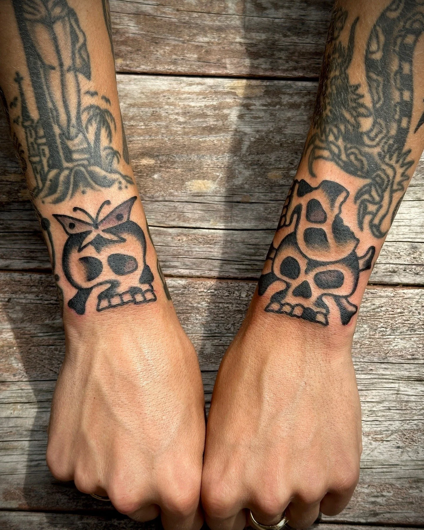 Two arms with tattoos of skulls and butterflies resting on a wooden surface.