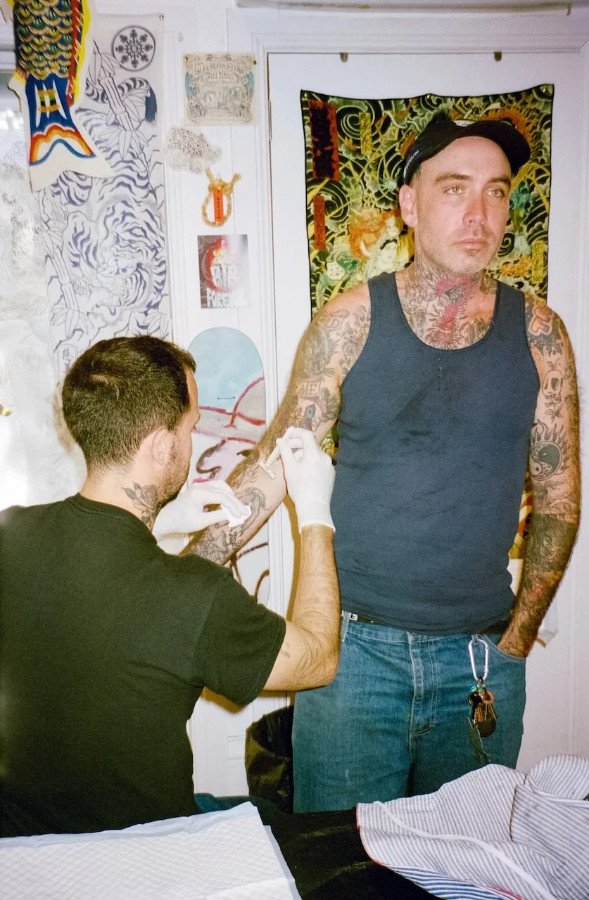 A man with tattoos on his arms and chest receives a tattoo on his right arm from a tattoo artist wearing gloves in a room decorated with artwork and posters.