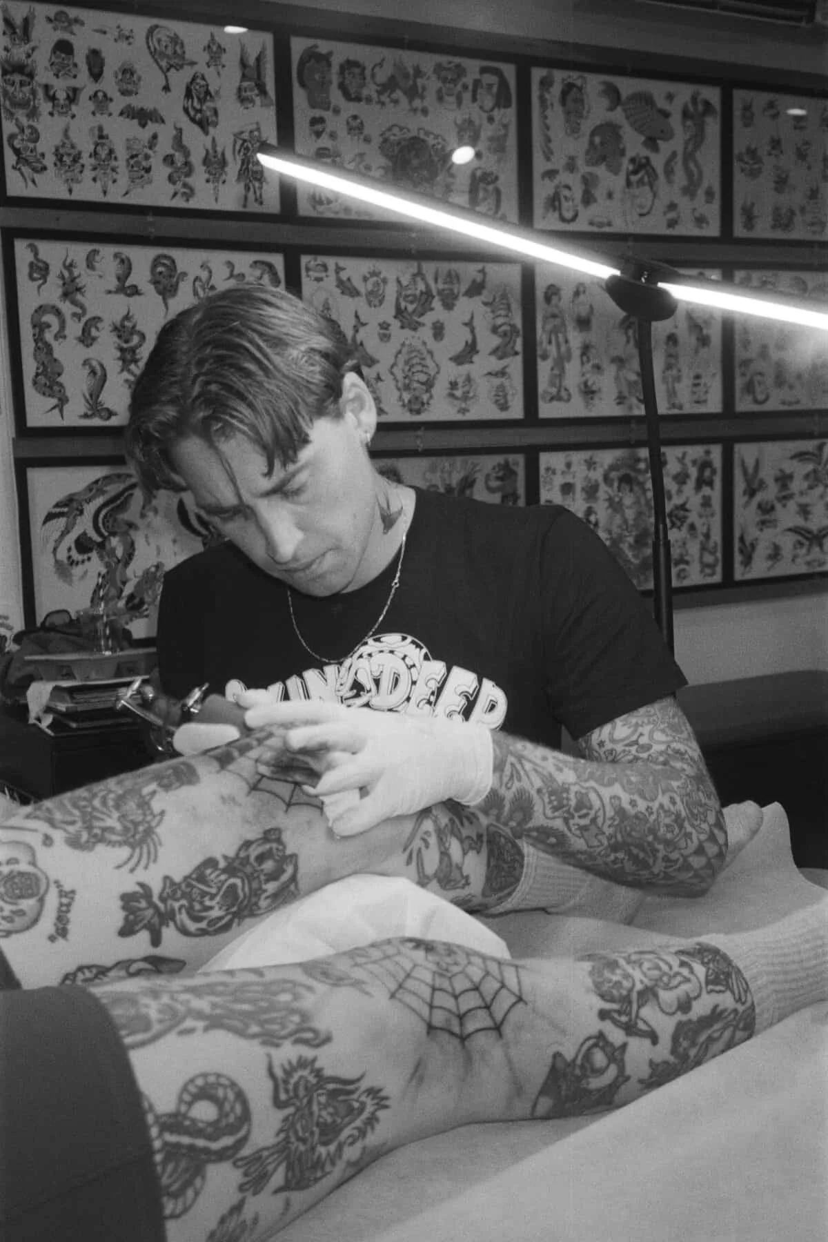 A tattoo artist working on a client's leg tattoo in a tattoo studio, with tattoo flash designs on the walls.
