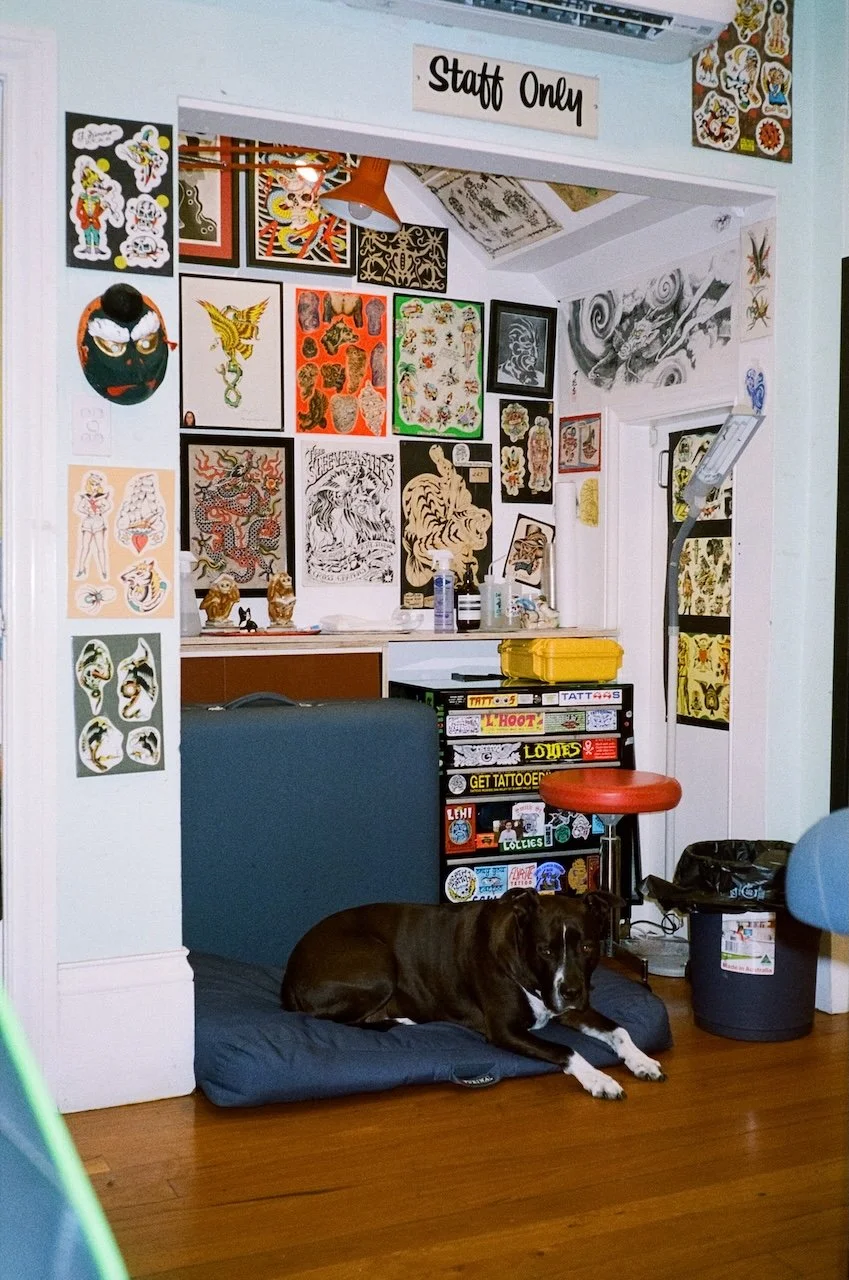 A tattoo studio with artwork on the walls, including traditional tattoo designs and sketches. There is a sign that reads 'Staff Only' above the doorway. In the foreground, a black and white dog is lying on a cushion on the wooden floor. The room appe