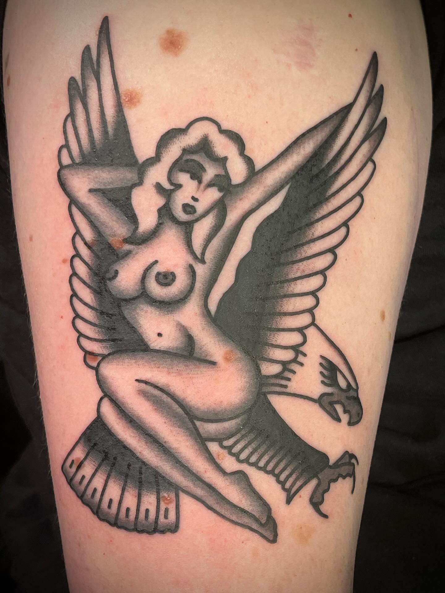 A black and gray tattoo of a nude woman with large wings, sitting on the tail of a bird with spread wings, on skin with some brown spots and redness.
