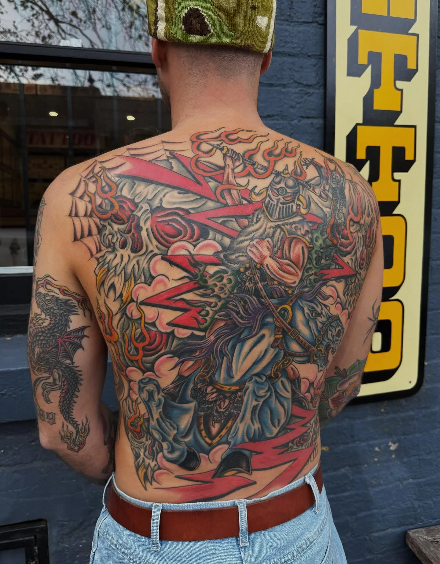 Colorful chest tattoo of a warrior with a helmet holding a weapon, surrounded by flames, skulls, clouds, and red lightning, with a dragon on the arm and a black background.