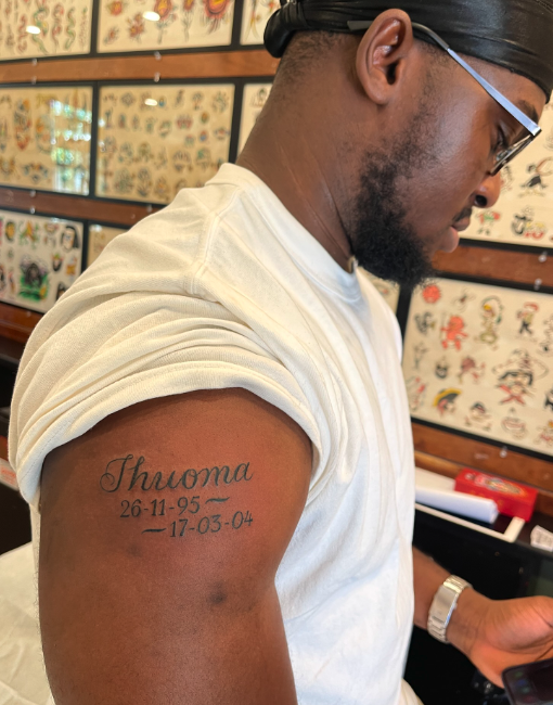 A man with glasses and tattoos on his upper arm reading "Thuyma," with dates 26-11-95 to 17-05-04, in a tattoo shop with framed tattoo designs on the wall.