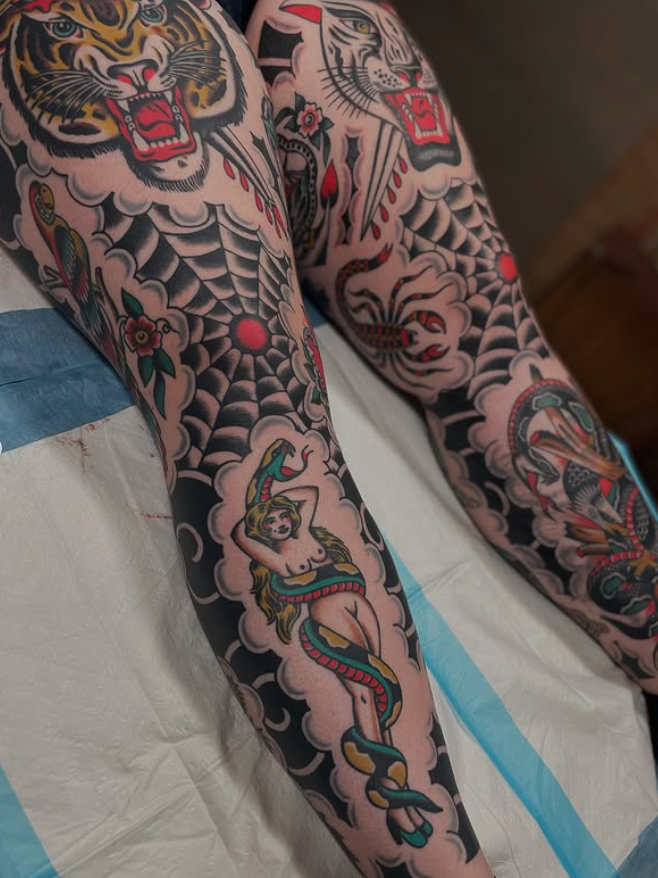 Tattooed arms with traditional American style tattoos, including a tiger, a snake wrapped around a woman, and a spider web.