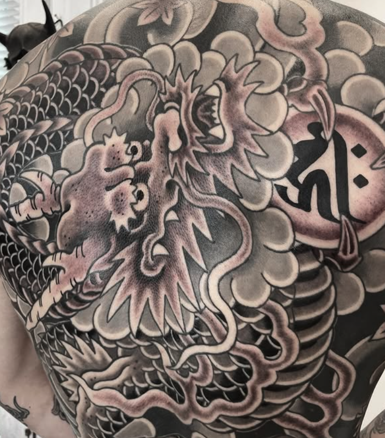 Black and gray tattoo of a fierce dragon with detailed scales, a prominent face, and swirling clouds surrounding it.