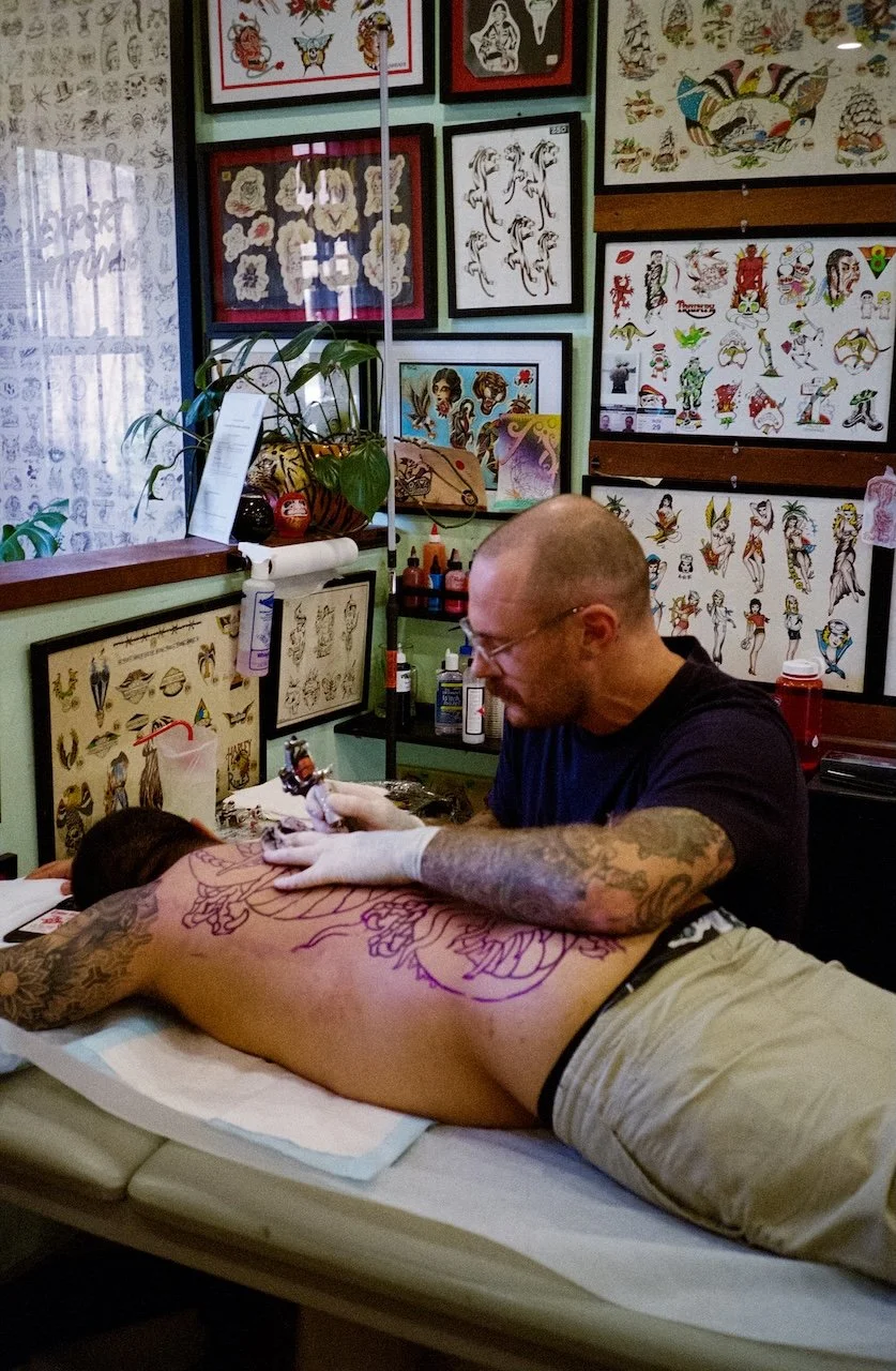 A man with tattoos tattooing a person's back in a tattoo studio decorated with tattoo flash and artwork on the walls.