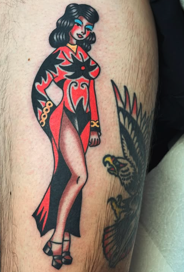 Tattoo of a vintage pin-up style woman with dark hair, colorful makeup, and a revealing red and black gown, standing next to a bird of prey.