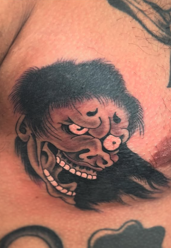 Tattoo of a stylized, traditional Japanese mask or face with exaggerated facial features, dark hair, and distinct black and pink colors.