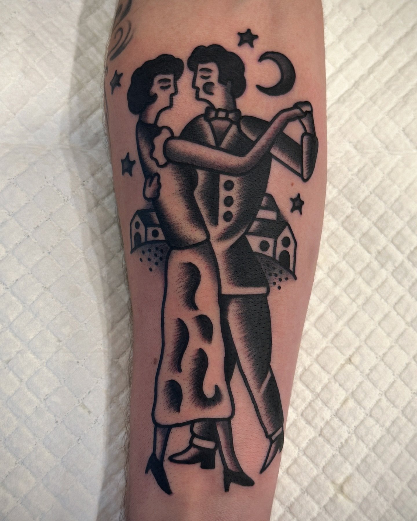 A black ink tattoo of a dancing couple in vintage style, with stars, a crescent moon, and houses surrounding them.